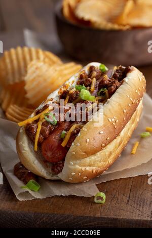 Chili Hot Dog mit Cheddar-Käse und grünen Zwiebeln auf Ein Sesambrötchen mit Kartoffelchips auf einem rustikalen Holzbrett Stockfoto