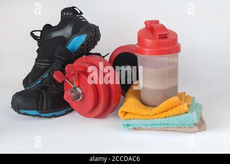 Fitness Lifestyle Konzept. Rote Hantel, bunte Handtücher, Shaker mit Schokoladenmolkenprotein und Sportschuhe. Isolierte Bodybuilding-Objekte Stockfoto