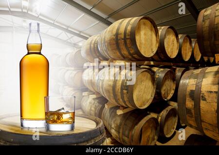 Eine Flasche und ein Glas bernsteinfarbener Whisky auf einem alten Fass in einem Fasslager, hinten beleuchtet mit Licht, das durch die Tür leuchtet. Stockfoto