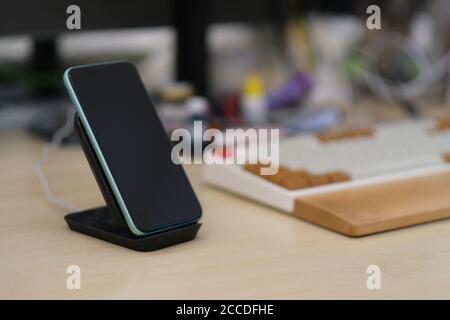 Nahaufnahme eines Smartphones mit vertikalem Ladegerät. Verschwommen Tastatur und Bürohintergrund Stockfoto