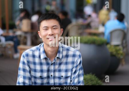 Porträt des jungen asiatischen Mannes in Casual Shirt im Café im Freien, lächelnd und Blick auf die Kamera. Hintergrund verwischen Stockfoto