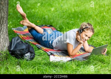 Junge Frau - beobachten pädagogische Webinar auf Laptop, sitzen auf grünem Gras im Stadtpark. Fernschulungskonzept. Stockfoto