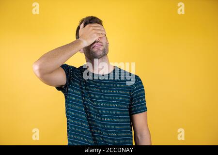 Junger schöner Mann bedeckt Gesicht mit einer Hand auf gelbem Hintergrund. Trägt ein blaues T-Shirt Stockfoto