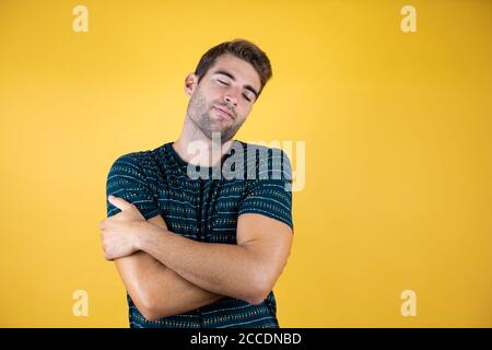 Junger gutaussehender Mann, der über isoliertem gelben Hintergrund steht und sich glücklich und positiv umarmt, selbstbewusst lächelt Stockfoto