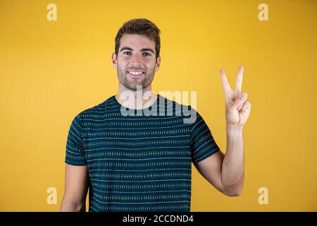 Jungen gutaussehenden Mann legere blaues T-Shirt über gelb isoliert Hintergrund lächelnd mit glücklichen Gesicht in die Kamera winking tun Sieg unterzeichnen. Anzahl Stockfoto