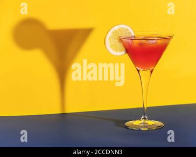 Roter Cocktail in einem Glas, das seinen Schatten mit einem formt Zitronenscheibe auf blauem und gelbem Hintergrund Stockfoto
