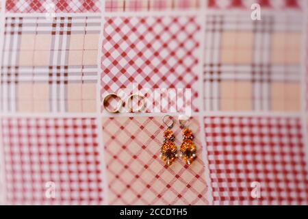 Budva, Montenegro - 21. august 2020: Nahaufnahme von Ohrringen mit Steinen und goldenen Eheringen auf kariertem Stoff. Stockfoto