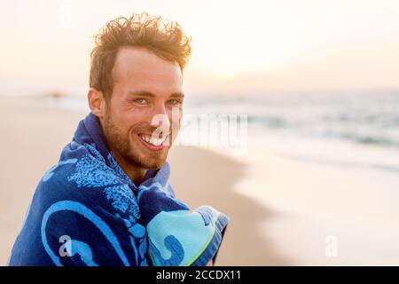 Lächelnd schöner junger Mann Schönheit Porträt am Strand bei Sonnenuntergang Blick auf Kamera lachen, gesundes Grinsen Gesicht glückliches Modell in Handtuch. Stockfoto