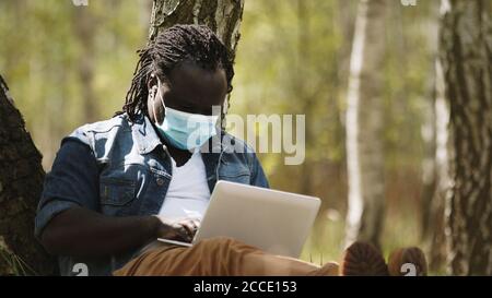 Remote-Work-Konzept. Afrikanischer Mann mit medizinischer Maske mit Laptop im Park. Hochwertige Fotos Stockfoto
