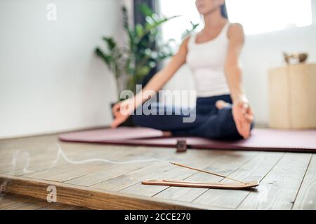 Zugeschnittenes Foto einer Frau, die in Lotusposition auf einer türkisfarbenen Yogamatte auf dem Boden meditiert. Konzentrieren Sie sich auf Räucherstäbchen und Rauch. Entspannen Sie sich nach dem Yoga-Training, h Stockfoto