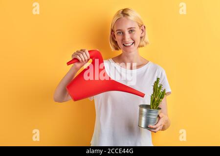 Schöne lächelnde positive Frau Bewässerung Blumen zu Hause, Pflanzenpflege. Nahaufnahme Foto. Isolierte gelbe Hintergrund, Studio erschossen. Lifestyle, Freizeit, Stockfoto
