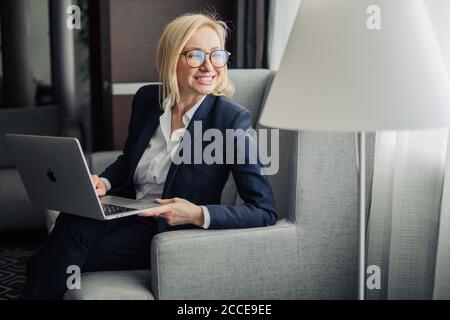 Attraktive blonde Wanderlehrerin in Brillen, trägt dunkelblauen Anzug und weißes Hemd arbeiten auf Laptop im Hotelzimmer. Reisen, Stockfoto