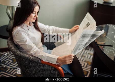 Porträt einer schönen Brünette Business Dame Zeitung lesen sitzen Im gemütlichen Sessel im Luxus-Büro Stockfoto
