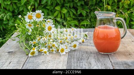 Grapefruitsaft in einem Krug und ein Bündel Gänseblümchen im Freien auf einem grünen unscharfen Hintergrund. Panorama. Stockfoto