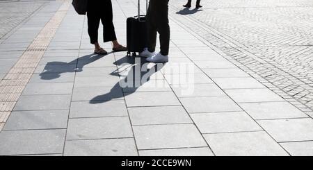 Nahaufnahme eines jungen Paares, das am Hauptbahnhof wartet Ein Koffer zwischen den Beinen und Schatten auf dem Bürgersteig Im Vordergrund Stockfoto