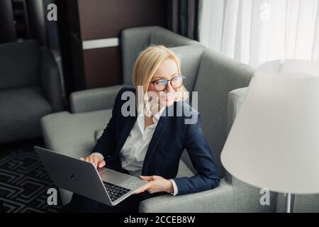 Attraktive blonde Wanderlehrerin in Brillen, trägt dunkelblauen Anzug und weißes Hemd arbeiten auf Laptop im Hotelzimmer. Reisen, Stockfoto