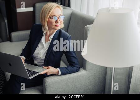 Attraktive blonde Wanderlehrerin in Brillen, trägt dunkelblauen Anzug und weißes Hemd arbeiten auf Laptop im Hotelzimmer. Reisen, Stockfoto