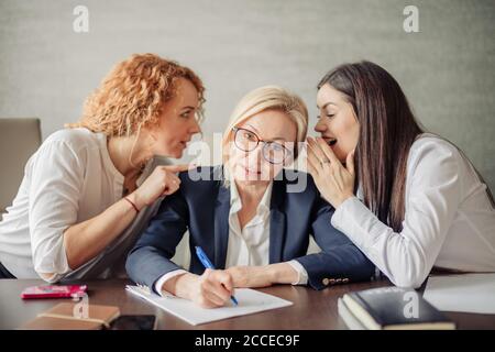 Zwei Bürotratschen flüstern von beiden Seiten in die Ohren der Chefin, erzählen ihr Gerüchte und stören Verleumdungen. Angewiderte, verärgerte Reaktion. Negativ Stockfoto