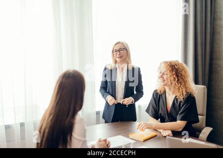 Weibliche Marketing-Experten sitzen um den Tisch und betrachten eine Sprecherin bei einem informellen Treffen. Blonde kaukasische weibliche Führungskraft, die eine Presentatio Stockfoto