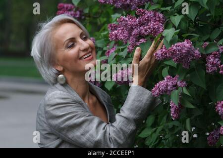 Reife blonde Frau in grauem Anzug geht im Garten, bewundert Blumen und lächelt, selektive Fokus Stockfoto