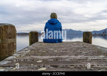 Europa, Deutschland, Bayern, Rosenheim, Prien am Chiemsee, Chiemsee, Mann sitzt auf dem Steg und blickt über den Chiemsee zu den Chiemgauer Alpen Stockfoto