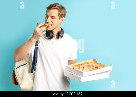 Porträt des jungen Mannes in weißem Hemd mit Rucksack Essen leckere Pizza Einnahme von Kurier, isoliert auf blauem Hintergrund Stockfoto