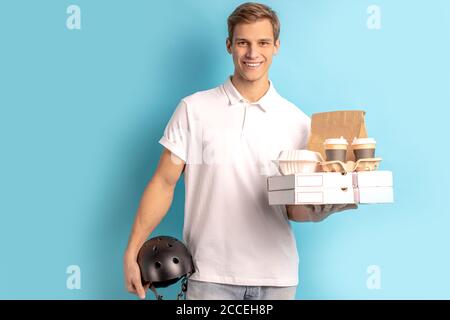 Porträt von schönen jungen kaukasischen Kerl hält Boxen mit Essen und Helm für Motorrad, trägt weißes T-Shirt, Lächeln und Blick auf Kamera isoliert o Stockfoto