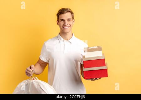 Portrait von jungen kaukasischen Kurier Mann mit Boxen und Kleidung für Kunden isoliert auf gelbem Hintergrund. Lieferservice Stockfoto