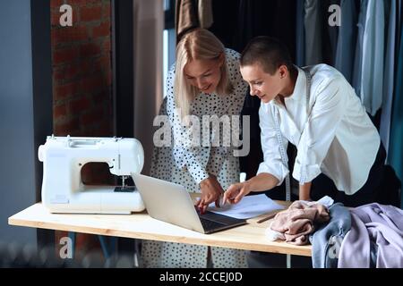 Schöne europäische Frau Blick auf Laptop, während in der Nähe von Tisch in der Werkstatt mit Kleidung, Laptop und Nähmaschine stehen. Fröhlich und glücklich zu schaffen Stockfoto