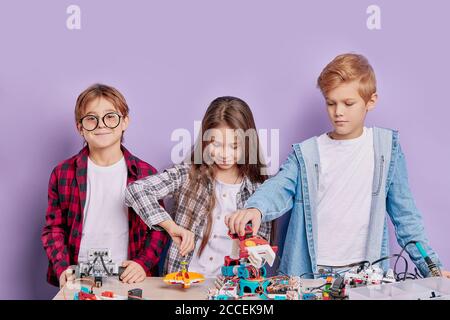 Portrait von begeisterten drei Kindern, die als Team arbeiten, Roboter montieren. Drei zukünftige Ingenieure schauen auf einen Tisch voller Robotik, zwei Jungen und ein Mädchen Stockfoto