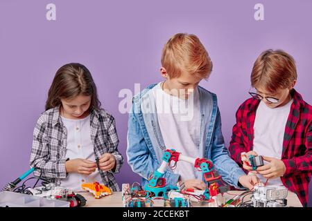 Portrait von begeisterten drei Kindern, die als Team arbeiten, Roboter montieren. Drei zukünftige Ingenieure schauen auf einen Tisch voller Robotik, zwei Jungen und ein Mädchen Stockfoto