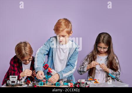 Portrait von begeisterten drei Kindern, die als Team arbeiten, Roboter montieren. Drei zukünftige Ingenieure schauen auf einen Tisch voller Robotik, zwei Jungen und ein Mädchen Stockfoto