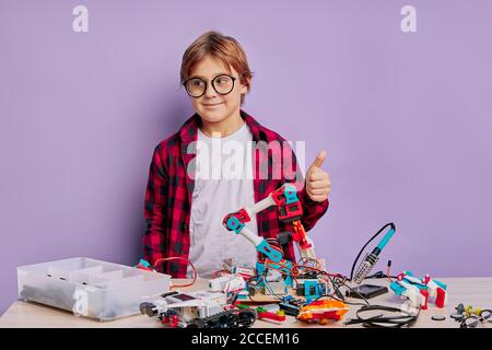 Kreatives Kind Junge trägt lässiges rotes Hemd mag Montage Roboter isoliert im Studio üben, Junge zeigen Daumen nach oben und Blick auf Kamera, lila Rücken Stockfoto