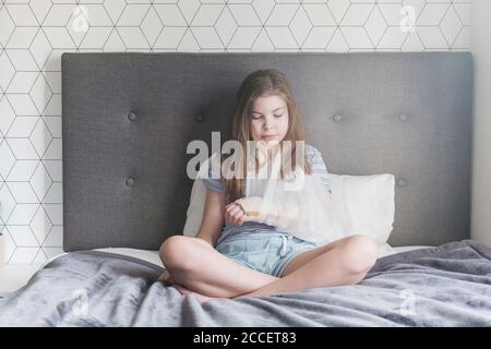 Mädchen sitzt auf dem Bett zu Hause Blick auf sie gebrochen Arm in einer Schlinge Stockfoto