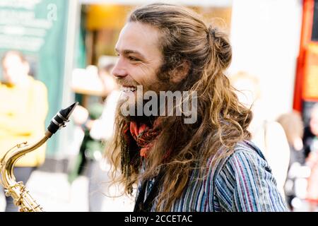 Porträt des Profils eines jungen Mannes mit lang Haare lächeln beim Saxophon spielen auf der Straße Stockfoto