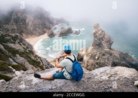 Erwachsener Rüde mit Rucksack ruht am Steinrand von Praia da Ursa Strand Küste. Surreale Landschaft neblige Landschaft von Sintra, Portugal. Stockfoto