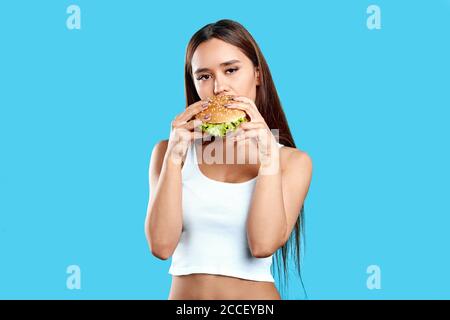 Junge schöne Frau in Mode weiß top Werbung lecker Burger. Nahaufnahme Foto. Frühstück, Snack, Geschäft, isoliert blauen Hintergrund Stockfoto