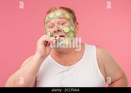 Junger lustiger übergewichtiger Mann mit schockiertem Gesicht, der skeptisch und sarkastisch aussagt und eine Scheibe Gurke aus der Maske auf seinem Gesicht testet, auf pinkfarbenem Rücken Stockfoto