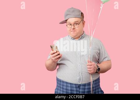 Lustige plump Kerl stehend hält Smartphone und Luftballons Zweifel an der Kamera Blick erwägen, ob zur Geburtstagsparty gehen, isoated auf pi Stockfoto