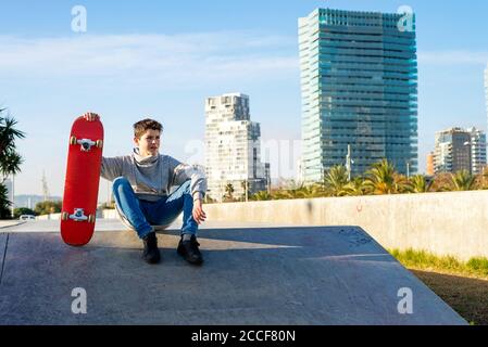 Kleiner Junge, der auf der Skatepark Rampe sitzt und ein Skateboard hält und wegschaut Stockfoto