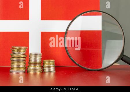 Vergrößerungsglas und Münzen vor der dänischen Flagge, Wirtschaftskonzept des Landes Stockfoto