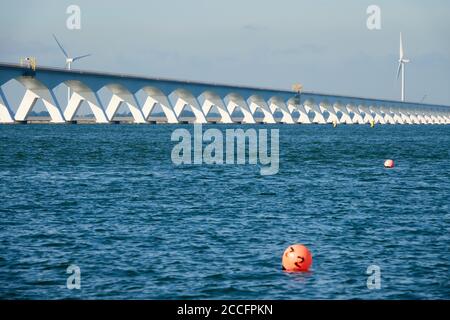 Lange Zeeland Brücke, tiefblaues Wasser mit roter Boje, Windturbine im Hintergrund. Niederlande, Zeeland. Stockfoto