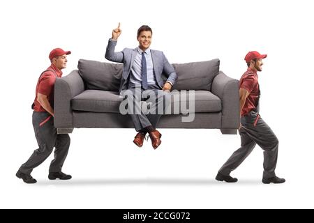 Lächelnder Mann in eleganten Kleidern sitzt auf einem Sofa und Zeigt nach oben, während zwei Mover das Sofa isoliert tragen Auf weißem Hintergrund Stockfoto