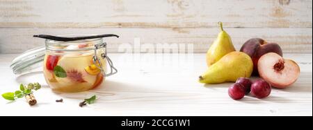 Birnen, Pfirsiche und Kirschpflaumen, frisch und in Dosen in einem Glas mit Zimt, Minze und Vanille auf weiß lackiertem Holzhintergrund, Panorama Stockfoto