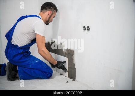 Der Arbeiter befestigt die Fliesen mit Zement an der Wand. Er trägt eine blaue Uniform und schwarze Handschuhe Stockfoto