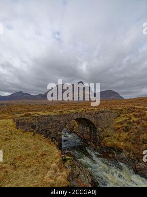 Schnell fließendes Wasser kommt durch einen Bogen der alten Stone Road Bridge, mit Bergen im Hintergrund an einem nassen Tag im April in den Highlands. Stockfoto