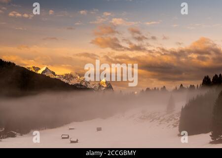 Zugspitze, Alpspitze und Wettersteingebirge am frühen Morgen. Im Vordergrund ein paar kleine Heuhaufen, Holzhütten und Nebel, während die Dämmerung baden Stockfoto