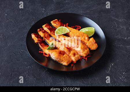 teriyaki Lachsfilet im Ofen gebacken, serviert auf einem schwarzen Teller mit Limettenkeilen auf einem Betontisch, horizontale Ansicht von oben Stockfoto