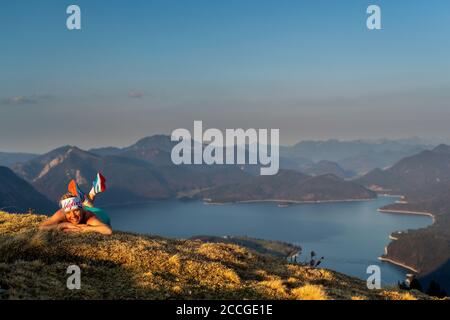 Trailrunnerin Katharina Kirschner liegt auf einer Frühlingswiese auf Simetsberg, im Hintergrund der Walchensee und lächelt die Kamera an. Stockfoto