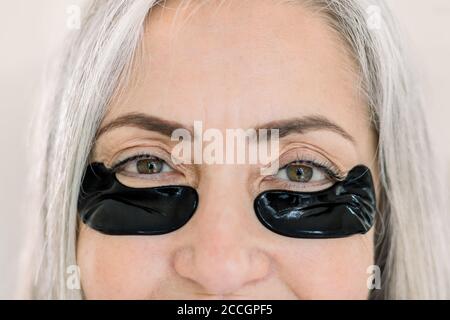 Nahaufnahme Schönheit beschnittenes Porträt von charmanten grauhaarigen Senior Dame, mit schwarzen unter dem Auge Hydrogel Patches, Blick auf die Kamera, während sie auf Stockfoto